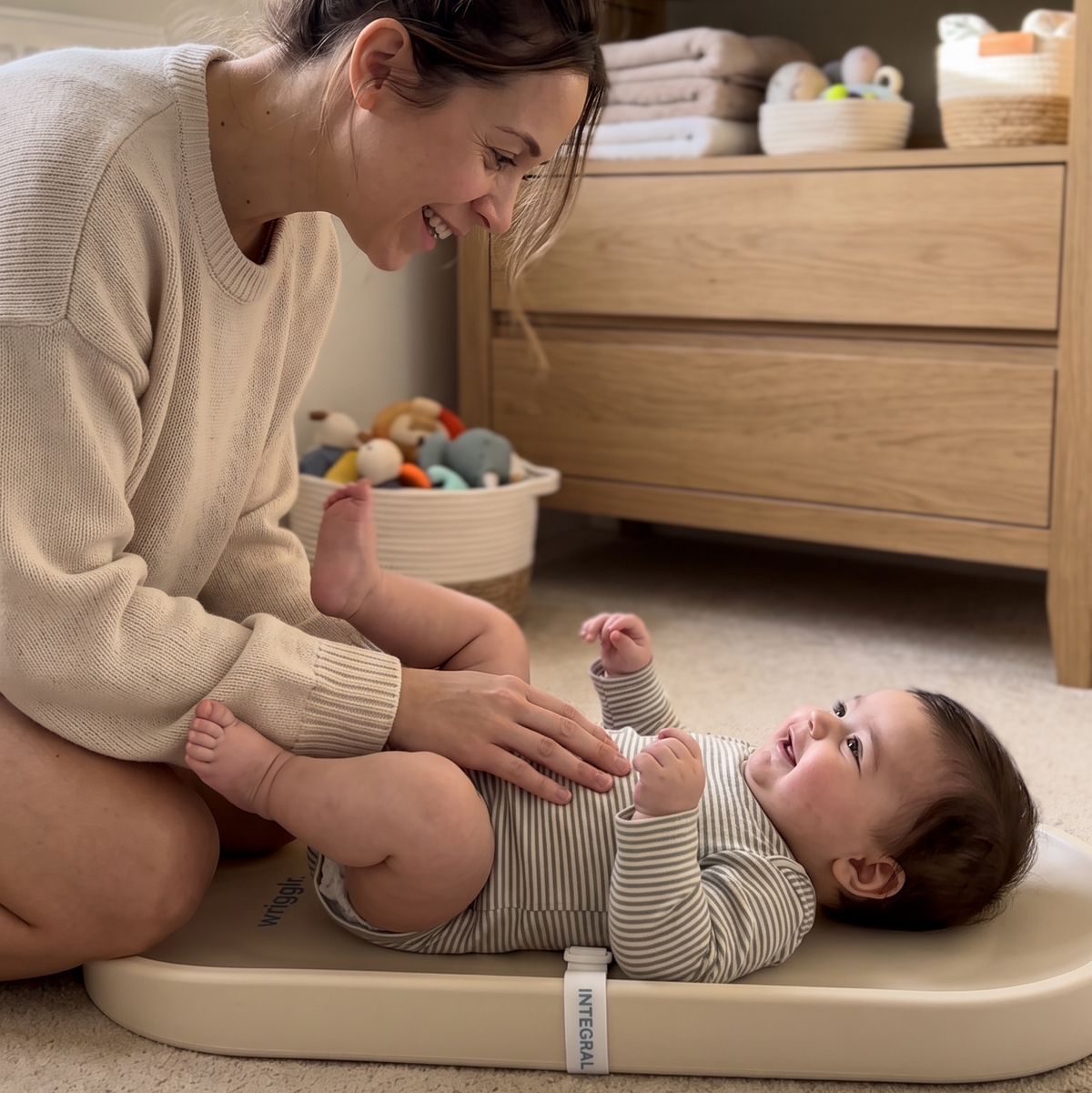 A baby in a striped outfit lies on a beige changing pad on a carpeted floor while a woman kneels beside them in a softly lit nursery. A wooden dresser and folded textiles are visible in the background.