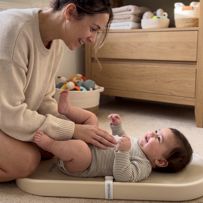 A baby in a striped outfit lies on a beige changing pad on a carpeted floor while a woman kneels beside them in a softly lit nursery. A wooden dresser and folded textiles are visible in the background.